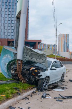Şehir yolundaki bir kazadan sonra ağır hasar görmüş beyaz bir sedan. Araba bir direğe çarptı, ön kısmı parçalandı, kaput açık. Kaza, yol tehlikesi ve sigorta kavramı.
