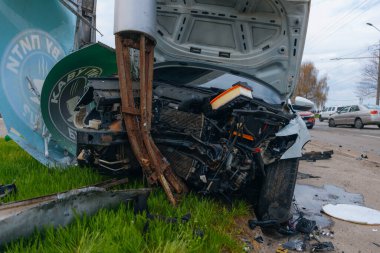 Şehir yolundaki bir kazadan sonra ağır hasar görmüş beyaz bir sedan. Araba bir direğe çarptı, ön kısmı parçalandı, kaput açık. Kaza, yol tehlikesi ve sigorta kavramı.