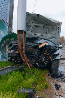 Şehir yolundaki bir kazadan sonra ağır hasar görmüş beyaz bir sedan. Araba bir direğe çarptı, ön kısmı parçalandı, kaput açık. Kaza, yol tehlikesi ve sigorta kavramı.