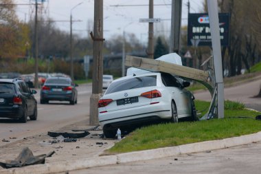 Şehir yolundaki bir kazadan sonra ağır hasar görmüş beyaz bir sedan. Araba bir direğe çarptı, ön kısmı parçalandı, kaput açık. Kaza, yol tehlikesi ve sigorta kavramı.