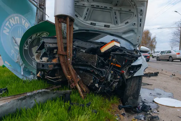 Şehir yolundaki bir kazadan sonra ağır hasar görmüş beyaz bir sedan. Araba bir direğe çarptı, ön kısmı parçalandı, kaput açık. Kaza, yol tehlikesi ve sigorta kavramı.