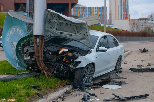 Şehir yolundaki bir kazadan sonra ağır hasar görmüş beyaz bir sedan. Araba bir direğe çarptı, ön kısmı parçalandı, kaput açık. Kaza, yol tehlikesi ve sigorta kavramı.
