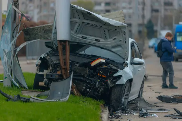 Şehir yolundaki bir kazadan sonra ağır hasar görmüş beyaz bir sedan. Araba bir direğe çarptı, ön kısmı parçalandı, kaput açık. Kaza, yol tehlikesi ve sigorta kavramı.