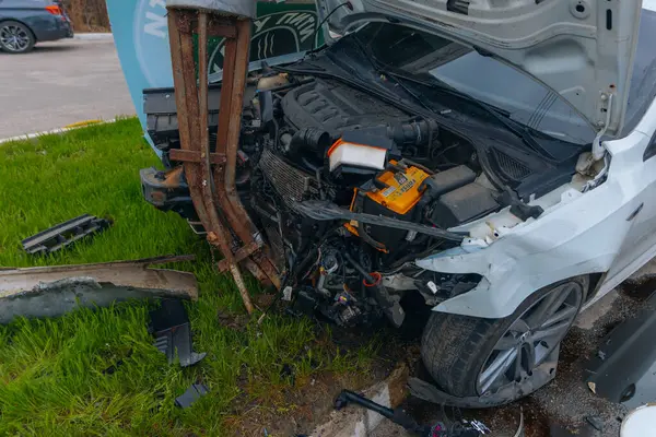 Şehir yolundaki bir kazadan sonra ağır hasar görmüş beyaz bir sedan. Araba bir direğe çarptı, ön kısmı parçalandı, kaput açık. Kaza, yol tehlikesi ve sigorta kavramı.