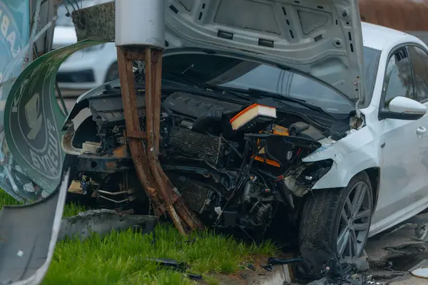 Şehir yolundaki bir kazadan sonra ağır hasar görmüş beyaz bir sedan. Araba bir direğe çarptı, ön kısmı parçalandı, kaput açık. Kaza, yol tehlikesi ve sigorta kavramı.
