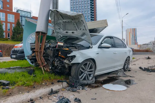 Şehir yolundaki bir kazadan sonra ağır hasar görmüş beyaz bir sedan. Araba bir direğe çarptı, ön kısmı parçalandı, kaput açık. Kaza, yol tehlikesi ve sigorta kavramı.