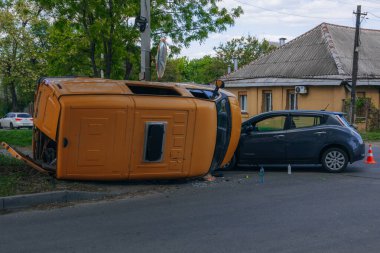 Bir şehir caddesinde iki arabalık bir kaza meydana geldi, bunlardan biri devrildi. Kırık farlar, yamuk bir kaput, kırık bir tampon ve cam kırıkları görülebilir.. 