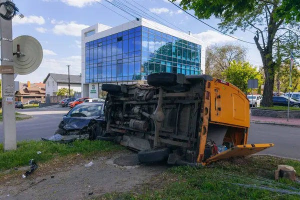 Bir şehir caddesinde iki arabalık bir kaza meydana geldi, bunlardan biri devrildi. Kırık farlar, yamuk bir kaput, kırık bir tampon ve cam kırıkları görülebilir.. 