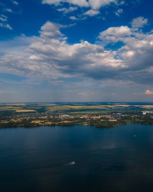 Büyük bir su kütlesi ve sahil şehri panoramik drone görüntüsü. Ön planda, küçük bir teknenin hareket ettiği koyu mavi bir su var. Dramatik kümülüs bulutları ve parlak mavi bir gökyüzü.