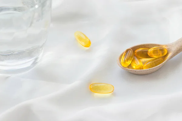 Pills of omega 3 in a spoon and a glass of water on white fabric background. Health care concept. Supplements and vitamins. Selective focus