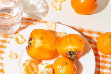 Creative layout made of fresh persimmon fruits, orange tangerin and a glass of water on cage kitchen towel. Healthy food concept. Summer picnic idea.