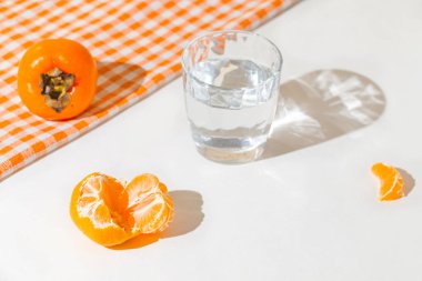 Creative layout made of fresh orange tangerin, persimmon fruit and a glass of water on cage kitchen towel and white background. Healthy food concept. Summer picnic idea.