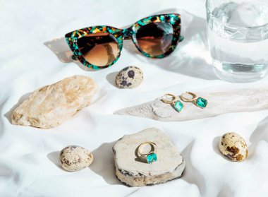 Güneş gözlüğü, taş, ahşap, bıldırcın yumurtası ve bir bardak suyla beyaz zemin üzerinde altın yüzük ve küpelerden oluşan yaratıcı bir kompozisyon. Ürün hala hayat konsepti. Seçici odak