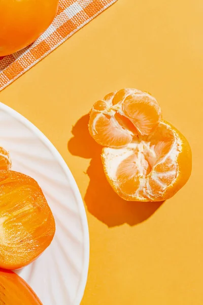 Creative layout made of orange mandarin closeup, fresh persimmon fruits on white plate on bright background with shadow. Healthy food concept. Summer picnic idea. Top view
