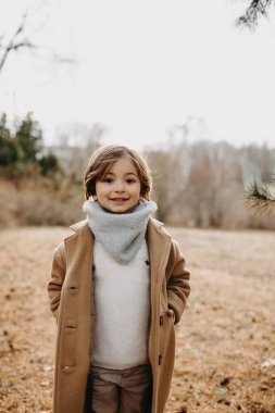 Sonbahar günü parkta yün ceket giyen küçük bir çocuk kameraya bakıyor ve gülümsüyor..