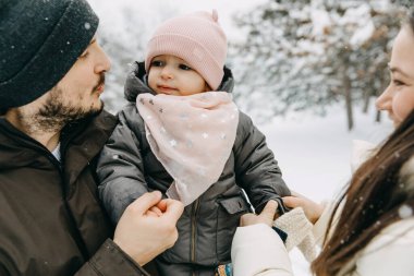 Karlı kış gününde üç kişilik bir ailenin yakın plan portresi. Baba ve anne küçük kızı kucaklıyor..