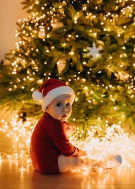 Noel Baba şapkalı bebek, Noel ağacında oturuyor. Işıklar arka planda, evde..