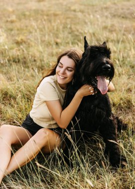 Genç esmer kadın tarlada oturuyor. Büyük siyah bir köpek, dev bir schnauzer cinsinin yanında..