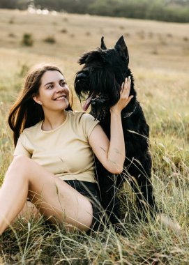 Genç esmer kadın kocaman siyah bir köpeğe sarılıp okşuyor, dev bir schnauzer cinsi..