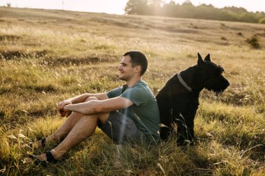 Genç adam büyük siyah bir köpek olan dev bir schnauzer cinsinin yanındaki tarlada oturuyor..