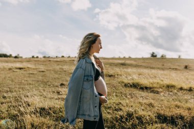 Kot ceket giyen sarışın hamile bir kadın, açık bir alanda duruyor, başka tarafa bakıyor..