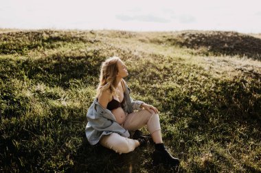 Kot ceket giyen sarışın hamile bir kadın, açık bir alanda oturuyor, başka tarafa bakıyor..