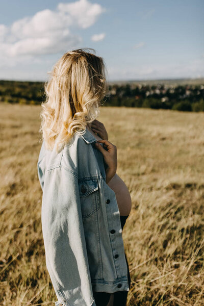 Blonde pregnant woman wearing a denim jacket, standing in an open field, looking away.