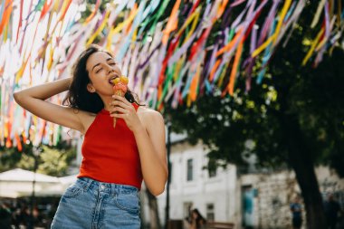 Happy young brunette woman walking on city street on a hot summer day, eating a double layered ice cream.