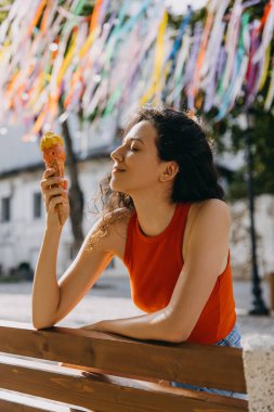 Happy young brunette woman sitting on a bench, eating ice cream, on hot summer day.