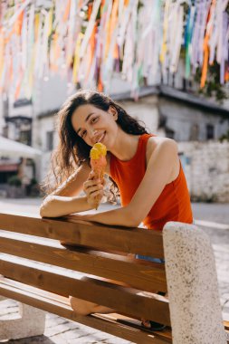 Happy young brunette woman sitting on a bench, eating ice cream, on hot summer day.