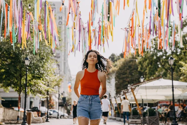Confident young woman with curly brunette hair, smiling, walking on city streets on hot summer day.