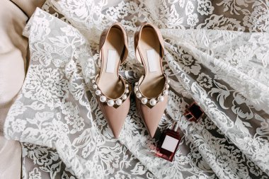 Pearl elegant wedding shoes and a bottle of perfume placed on a lace bridal dress, top view.