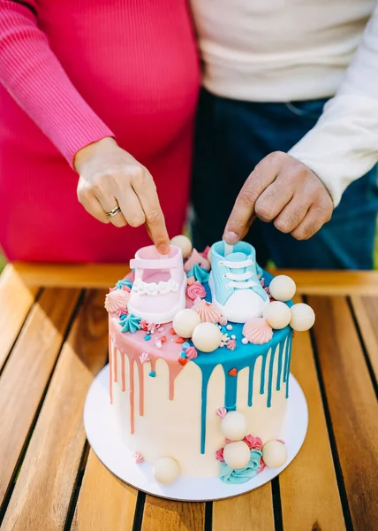 Closeup of man and woman with a gender reveal party cake in pink and blue colors with small baby shoes on top.