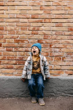 Toddler boy yelling outdoors sitting by a brick wall, on city streets.