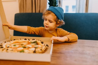 Little boy sitting at a cafe table opening a pizza box with traditional margherita pizza.