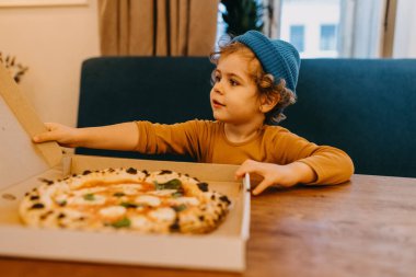 Little boy sitting at a cafe table opening a pizza box with traditional margherita pizza.