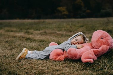 Little child wearing a bunny costume, laying on a big pink plush bunny toy, in an open field.