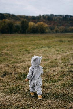 Little child in a plush mouse costume walking in an open field towards a forest.