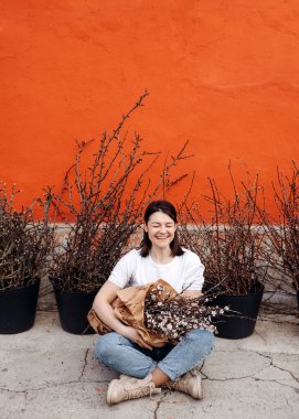 Kadın gülüyor, yerde oturuyor, elinde pembe kiraz çiçekleriyle bir sürü dal tutuyor..