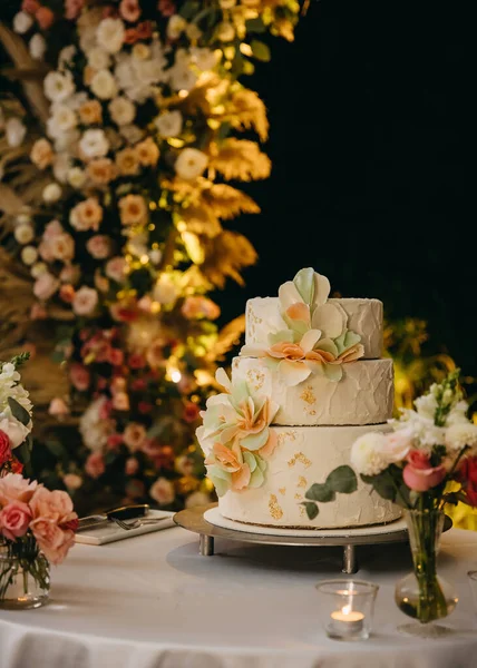 Three layered wedding cake at night decorated with flowers made of frosting.