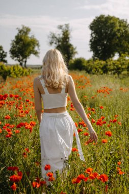 Beyaz elbiseli, vahşi kırmızı gelinciklerle tarlada yürüyen sarışın bir kadın..