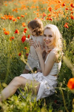 Genç anne ve küçük oğlu vahşi kırmızı gelinciklerle bir tarlada oturup oynaşıyor ve sarılıyorlar..