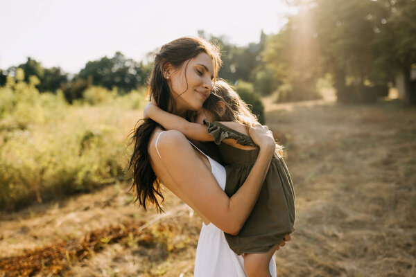 Mother holding her little daughter in arms, smiling, hugging, outdoors in a park.