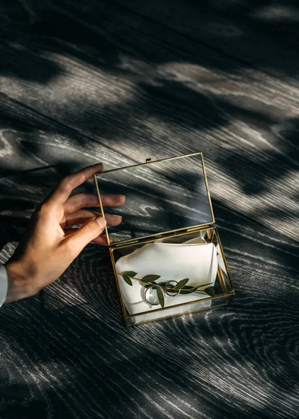 An elegant hand opens a glass box revealing wedding rings on a silk cloth with light casting dramatic shadows on a wooden surface.