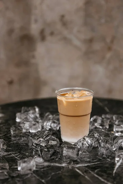 An iced coffee in a plastic cup surrounded by scattered ice cubes on a marble surface.