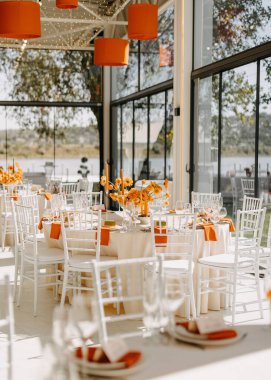 Beyaz sandalyeleri, zarif masa düzenleri ve turuncu renkli doğal ışık süslemeleri olan güzel bir etkinlik alanı..