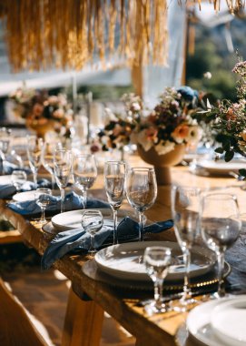 Rafine cam, beyaz tabak ve düzgünce katlanmış mavi peçetelerle süslenmiş uzun ahşap bir yemek masası sunan zarif bir köy düğünü.. 