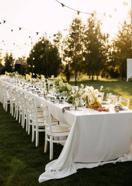 Kırsal ahşap sandalyeli şık bir açık hava sofrası, gün batımında sicim ışıkları altında yeşillik, üzüm ve çiçeklerle süslenmiş uzun beyaz bir masa..