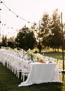 Kırsal ahşap sandalyeli şık bir açık hava sofrası, gün batımında sicim ışıkları altında yeşillik, üzüm ve çiçeklerle süslenmiş uzun beyaz bir masa..