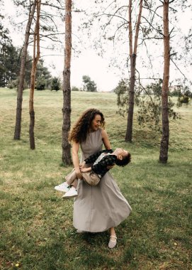 Annesi gülen oğlunu ağaçlarla çevrili bir parkta sallıyor..